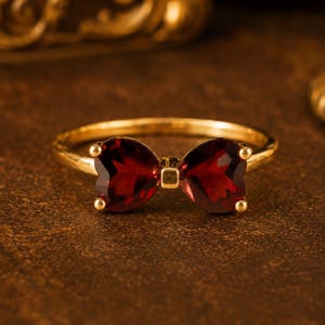 May include: A gold ring featuring a bow design composed of two deep red, heart-shaped faceted gemstones. The center of the bow is accented with a small gold square, and the band is a smooth, polished gold metal, creating a cohesive piece of jewelry.