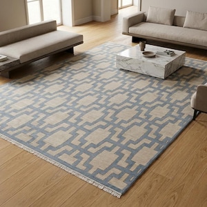 Puede incluir: Una alfombra rectangular con un patrón geométrico repetitivo en tonos azul apagado y beige. La alfombra tiene una textura de pelo corto y está terminada con flecos blancos cortos a lo largo de los dos bordes más cortos, sobre un suelo de madera.