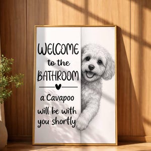 Op de afbeelding: Een rechthoekige wandprint met een witte achtergrond en een zwart-witte illustratie van een pluizige Cavapoo-hond. De tekst luidt: Welcome to the BATHROOM a Cavapoo will be with you shortly. De print is ingelijst in een dunne gouden rand.