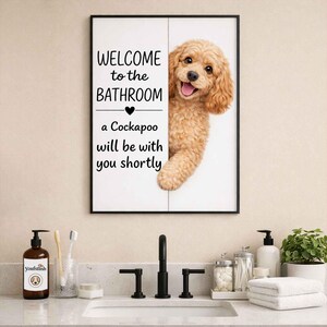 Pode incluir: Uma placa de casa de banho emoldurada com o texto "WELCOME to the BATHROOM" e "a Cockapoo will be with you shortly". Um cão Cockapoo espreita por trás da placa. A placa está numa casa de banho.