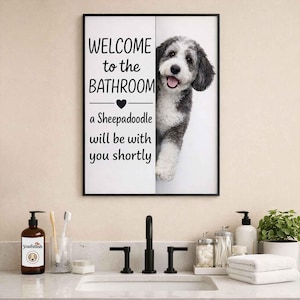 May include: Framed bathroom wall art with a black border. The artwork features the text "WELCOME to the BATHROOM" above a heart and the phrase "a Sheepadoodle will be with you shortly." A Sheepadoodle dog peeks from behind the artwork.