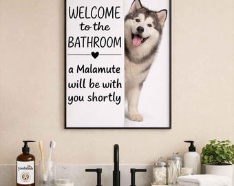 Könnte beinhalten: Gerahmtes Badezimmerschild mit dem Text "WELCOME to the BATHROOM" und "a Malamute will be with you shortly." Ein lächelnder Malamute-Hund schaut hinter dem Schild hervor. Das Schild befindet sich in einem Badezimmer mit Waschbecken und Zubehör.