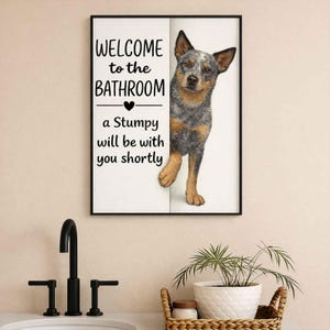 Può includere: Arte da parete per bagno con cornice nera. L'arte presenta un cane da pastore australiano blu che sbuca da dietro uno sfondo bianco. Il testo recita "WELCOME to the BATHROOM" e "a Stumpy will be with you shortly."