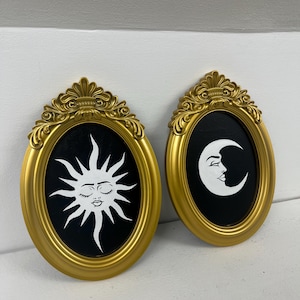 May include: Two gold-coloured, oval-shaped wall art pieces. One depicts a white sun with closed eyes on a black background. The other shows a white crescent moon with a face on a black background. The frames have decorative detailing.