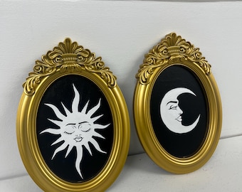 May include: Two gold-framed oval wall art pieces. One features a white sun with closed eyes on a black background. The other displays a white crescent moon with a face on a black background. The frames have ornate detailing.