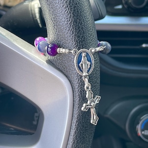 May include: A rosary with purple and blue beads hangs from a steering wheel. The rosary features a silver crucifix and a pendant with a blue oval and a depiction of the Virgin Mary.