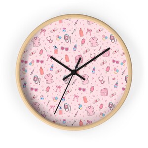 May include: A round wall clock with a light wood frame. The clock face is pink and covered in a repeating pattern of medical-themed illustrations, including scrubs, stethoscopes, and bandages. Black clock hands indicate the time.