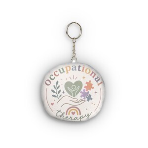 May include: A round keyring featuring a white background with the text occupational therapy in a pastel rainbow gradient. The design includes a central heart with a figure, a hand, a leafy branch, puzzle pieces, and a small rainbow icon.