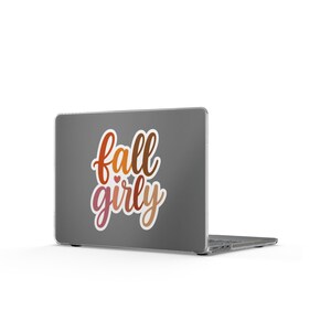 May include: A dark gray laptop featuring a sticker with the text fall girly in a cursive font. The lettering displays a gradient of orange, red, and brown tones, with a small heart icon positioned between the two words on the laptop lid.
