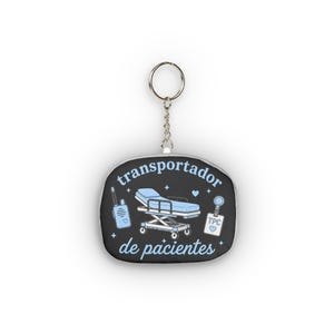 May include: A black keychain with a silver metal ring featuring a light blue illustration of a hospital gurney, a walkie-talkie, and an ID badge. The text "transportador de pacientes" is printed in light blue, surrounded by small star and heart icons.