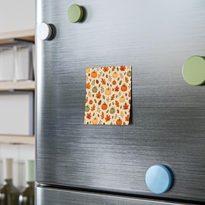 May include: A square magnet featuring a repeating pattern of orange and green pumpkins, brown acorns, and autumn leaves in shades of red, orange, and yellow on a cream background, attached to a brushed stainless steel refrigerator door.