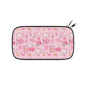 Könnte beinhalten: Rosa Federtasche mit schwarzem Reißverschluss und Anhänger. Das Muster zeigt medizinische Geräte wie Stethoskope, Spritzen und Medikamentenflaschen. Das Design ist in Rosa, Weiß und Orange gehalten.