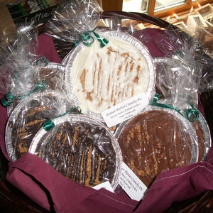 May include: A wicker basket filled with several wrapped peanut butter crunchy pies. The pies are in aluminum foil tins, covered in clear plastic, and tied with green ribbon. The pies have a variety of toppings, including white icing and chocolate drizzle.