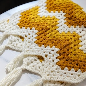 May include: A handmade crochet blanket in a granny square pattern. The blanket features a color block design with white and yellow squares. White tassels are attached to the corners of the blanket. The blanket is made of soft yarn.