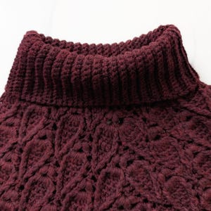 May include: Close-up of a burgundy turtleneck sweater. The sweater features a ribbed turtleneck and a textured, patterned knit design. The color is a deep, rich shade of red-purple.