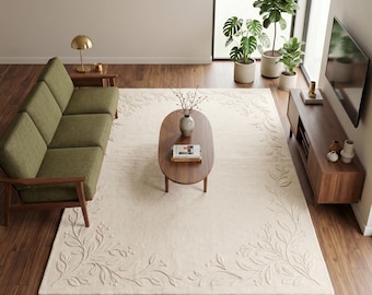 Könnte beinhalten: Ein großer rechteckiger cremefarbener Teppich mit einem geprägten floralen Rankenmuster entlang der Ränder. Der Teppich liegt in einem Wohnzimmer mit einem grünen Sofa, einem ovalen Couchtisch aus Holz und einem Holz-TV-Board auf Parkett.