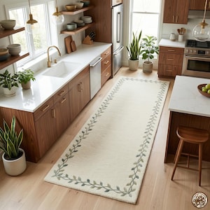May include: A long, rectangular runner rug featuring a cream-coloured centre and a decorative border of green, leafy vines. The rug is positioned on a light-coloured wooden floor in a kitchen with walnut cabinets, white countertops, and stainless steel appliances.