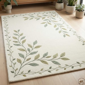 May include: Cream-colored rectangular rug with a botanical design. The rug features a border and corner accents of green and olive-colored leaves and vines. The rug is placed on a light-colored wooden floor.
