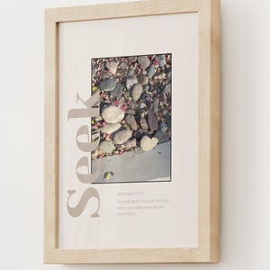 May include: Framed art print featuring a photograph of smooth stones and pink flower petals. The word "Seek" is printed vertically in a light brown color. The text "Jeremiah 29:13" and a quote are printed below the photograph.