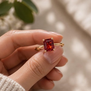 Può includere: Un anello in oro con una pietra preziosa rosso vibrante dal taglio rettangolare a smeraldo, fissata da quattro griffe. La fascia è sottile e lucida, con una finitura metallica brillante che esalta il colore profondo e saturo della pietra.