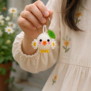 May include: A beaded keychain shaped like a small white bird with black eyes, an orange beak, pink cheeks, and yellow feet. The bird features small yellow wing accents and a single green leaf-shaped bead attached to a silver metal keyring.