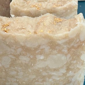 May include: Close-up of two bars of handmade soap. The soap is a creamy off-white color with a textured surface and gold flecks. The soap appears to be freshly cut, with a slightly rough edge. The overall aesthetic is natural and luxurious.