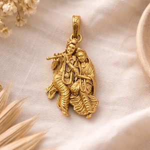 Könnte beinhalten: Goldfarbener Anhänger mit detaillierter Darstellung von zwei Figuren, von denen eine Flöte spielt. Die Figuren sind mit aufwendiger Kleidung und Schmuck geschmückt. Der Anhänger ist an einer Schlaufe oben aufgehängt, was darauf hindeutet, dass er als Halskette getragen werden soll.