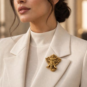 May include: A gold-toned metal brooch shaped like an elephant head with intricate filigree detailing and curved tusks, pinned to the lapel of a cream-coloured blazer worn over a matching high-neck top, showcasing a detailed metallic texture.
