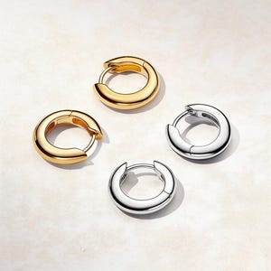 May include: Four hoop earrings, two in gold and two in silver, are displayed on a light surface. The earrings feature a simple, rounded design with a hinged clasp. The gold earrings have a polished finish, while the silver earrings have a shiny appearance.