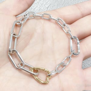 May include: Silver chain bracelet with a gold heart-shaped clasp. The bracelet features a series of rectangular links connected together. The clasp is a contrasting gold color, adding a touch of elegance to the piece. The bracelet is designed to be worn around the wrist.