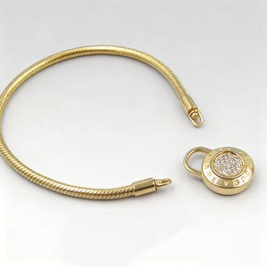 May include: A gold-coloured snake chain bracelet with a round, lock-shaped clasp. The clasp features small, clear stones and engraved text. The bracelet is unclasped, showing the two ends ready to be connected.