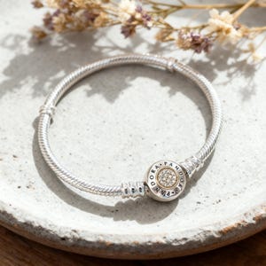 May include: A silver bracelet with a round clasp featuring the word "Pandora" and a circle of small, clear stones. The bracelet is on a light gray surface with dried flowers in the background.