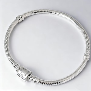 May include: A silver bracelet with a snake chain design. The bracelet features a cylindrical clasp with the brand name engraved on it. The bracelet is made of a shiny, reflective metal and is displayed on a white background.