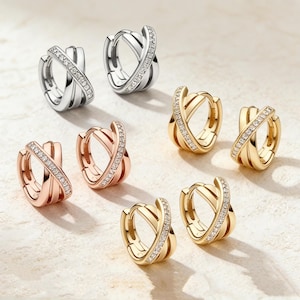 May include: An assortment of hoop earrings in silver, rose gold, and gold tones. Each earring features a crisscross design, with one band encrusted with small, sparkling gemstones. The earrings are displayed on a light-colored surface.