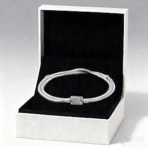 May include: A silver bracelet with a decorative clasp, displayed in an open white jewellery box with a black interior. The bracelet is made of a woven, textured material.
