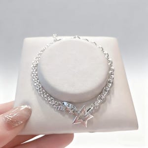 May include: A silver bracelet with a star-shaped charm. The bracelet features a double chain design and a clasp with small, clear stones. The star charm has a pinkish-silver hue and is outlined with small stones. The bracelet is displayed on a white surface.