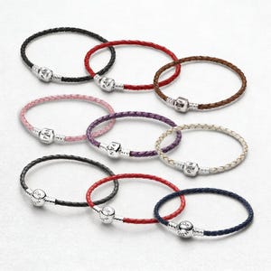 Peut inclure: Collection de bracelets en cuir tressé de différentes couleurs : noir, rouge, rose, violet, marron, blanc et bleu. Chaque bracelet est doté d'un fermoir argenté portant le nom de la marque. Présentation sur fond blanc.