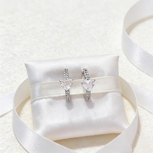 May include: A pair of silver hoop earrings with heart-shaped clear gemstones. The earrings are displayed on a white satin pillow with a white ribbon. The earrings have a delicate, elegant design.
