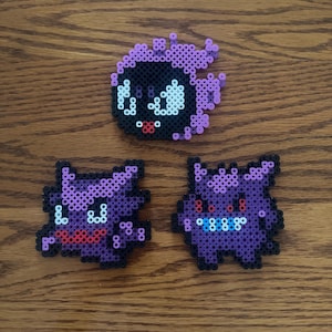 May include: Three pixel art figures of Pokemon characters. The top figure is black with purple accents. The bottom two figures are purple with black accents and red and blue details. The figures are made of small, colorful beads.