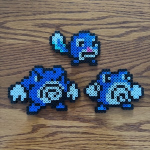May include: Three pixel art figures in blue, white, and black beads are arranged on a wooden surface. The figures appear to be stylized creatures, with one standing and two in a different pose. The figures are made with small, colorful beads.