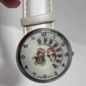 Op de afbeelding: Zilverkleurig polshorloge met een witte wijzerplaat met cartoonpinguïns met kerstmutsen. Het horloge heeft een witte leren band en zwarte cijfers. De woorden "JAPAN MOVT" staan op de wijzerplaat.