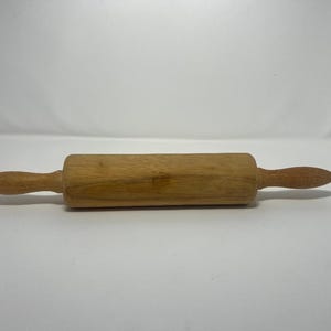 May include: A wooden rolling pin with a light brown finish. The rolling pin has a cylindrical body and two rounded handles. A small metal hook is attached to one handle. This kitchen tool is used for rolling out dough.