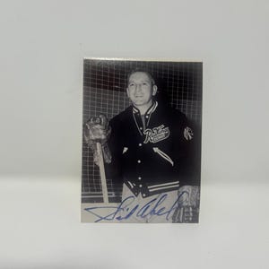 May include: A black and white vintage sports trading card featuring a hockey player wearing a Detroit Red Wings jacket with a team logo on the chest. The player holds a hockey stick and wears a glove. A blue ink autograph is signed across the bottom.