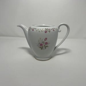 Puede incluir: Tetera de porcelana blanca con boquilla y asa curvadas. La tetera está decorada con motivos florales de rosas rosas y un borde dorado. El diseño incluye un pequeño grupo de rosas rosas y hojas verdes.