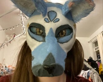 May include: A white and blue furry mask with large, upright ears. The mask features a blue snout and accents, with mesh eye coverings and star details. The design resembles a stylized canine or similar animal.