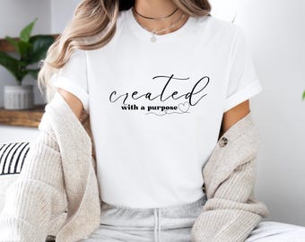 Puede incluir: Camiseta blanca con la frase "created with a purpose" en fuente cursiva negra. El diseño incluye un símbolo de corazón. La modelo lleva una rebeca beige y un pantalón de chándal gris. La camiseta tiene cuello redondo y mangas cortas.