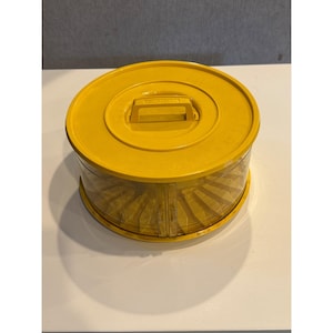 May include: A yellow plastic container with a handle on the lid. The container has a clear, ribbed, cylindrical body. The lid and handle are a solid yellow color. The container is sitting on a white surface.