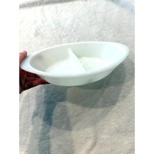 May include: A white oval-shaped serving dish made of milk glass featuring a single central divider that separates the interior into two distinct sections. The dish has smooth, curved edges and integrated handles on both sides for easy gripping.