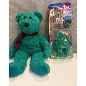 May include: A plush green bear with a red and black plaid scarf, next to a packaged green bear with a four-leaf clover. The packaging reads "ERIN THE BEAR" and features a castle illustration.