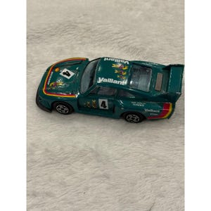 May include: A green toy race car with the number 4 on the side and hood. The car has the word "Vaillant" printed on the side and top, with colorful accents. The car is a miniature replica of a vintage race car.